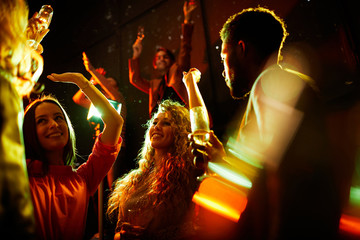 Cheerful pretty young ladies dancing with raised arms and talking to handsome men while flirting...