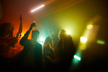 Group of carefree young multiethnic people dancing in disco lights and drinking alcohol at night...