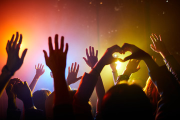 Crowd of unrecognizable fans waving hands and enjoying performance at concert, man making heart...