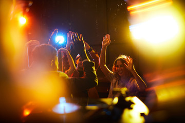 Cheerful excited young people dancing with raised hands at rocking party, blurred lights effect