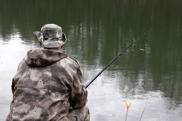 Fisherman in camouflage clothes sitting with a fishing rod on the shore. Man angling on the lake in...