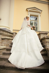Naklejka premium Beautiful Bride in a white Wedding Dress outdoors with a Bouquet of flowers and a veil on stairs