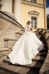 Fototapeta premium Beautiful Bride in a white Wedding Dress outdoors with a Bouquet of flowers and a veil on stairs