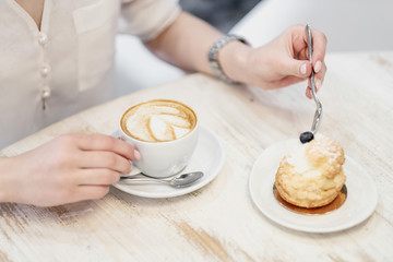 Cappuccino coffee with cup with crema in a heart wolf and cream cake dessert on a white wooden table in a cafe. Female hands with a spoon.