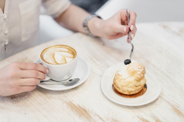 Cappuccino coffee with cup with crema in a heart wolf and cream cake dessert on a white wooden table in a cafe. Female hands with a spoon.