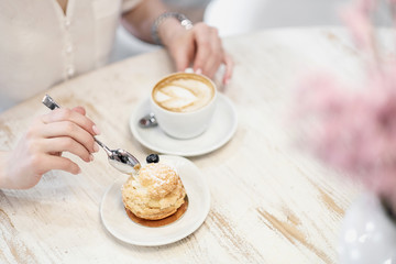 Cappuccino coffee with cup with crema in a heart wolf and cream cake dessert on a white wooden table in a cafe. Female hands with a spoon.