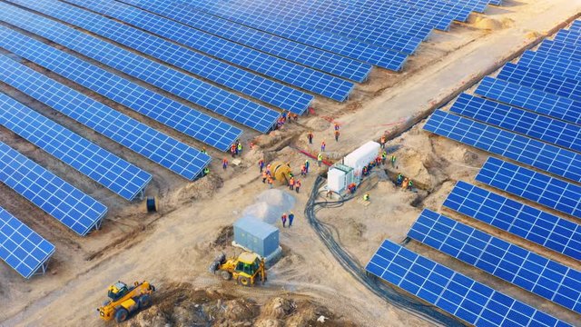 Aerial circling view of a giant industrial solar power station (solar electricity plant) with a group of workers installing a power cable in a transformer box 