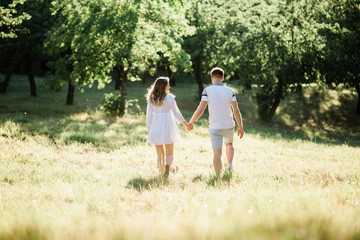 Back view of young pretty couple in love walking  in park. Handsome cheerful blonde girl in white dress hugging her boyfriend. Man and woman having fun outdoors