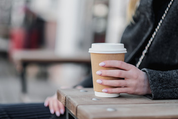 Close-up of woman's hands with a brown paper cup of coffee on an outdoor cafe bench. Uses a smartphone and the Internet.