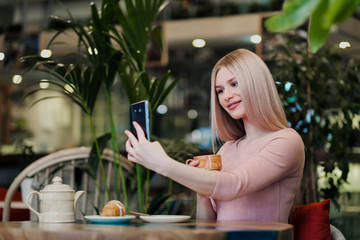 Young beautiful girl sitting in a green cafe at a table. Drinking tea with croissants, chatting, laughing and taking pictures on a smartphone.