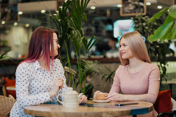 Two young beautiful girls sit in a green cafe at a table. Drink tea with croissants, chatting, laughing and taking pictures on a smartphone.