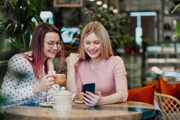 Two young beautiful girls sit in a green cafe at a table. Drink tea with croissants, chatting, laughing and taking pictures on a smartphone.