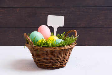 Colorful pastel eggs in basket with natural green grass with white label. Easter decoration concept on white concrete and wooden background