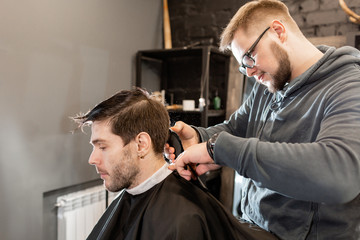 Barber work with clipper machine in barbershop. Professional trimmer tool cuts beard and hair on young guy in barber shop salon.
