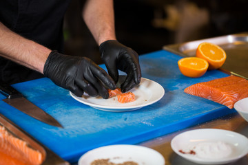 Theme cooking is a profession of cooking. Close-up of a Caucasian man's hand in a restaurant kitchen preparing red fish fillets salmon meat in black latex gloves uniform