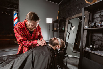 Beard cutting, face care. Barber work with clipper machine in barbershop. Professional trimmer tool cuts beard and hair of young guy in barber shop salon.