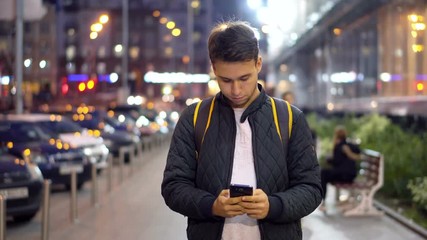 Young Attractive Man Typing Message Using Smartphone in the evening city street with bokeh light effect.