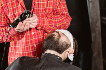 Naklejka premium Barber work with clipper machine in barbershop. Professional trimmer tool cuts beard and hair of young guy in barber shop salon.