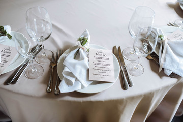 Wedding table setting. White plates, glasses, cutlery and napkins lie on the dinner round table decorated with greenery