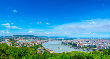 Naklejka na ściany i meble Panoramic cityscape view of hungarian capital city of Budapest from the Gellert Hill. The bridges connecting Buda and Pest across the river Danube. Summertime sunshine day, blue sky and green of trees