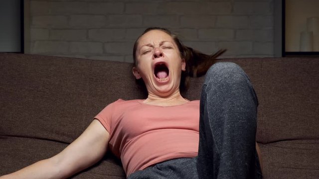 Sleepy woman yawns sitting on the couch watching TV at home. Close up.