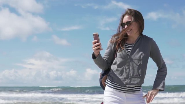 A woman takes a selfie photo on the phone on the background of the sea. A young woman takes herself on the phone. She is walking along the deserted beach