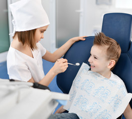 Kids girl and boy play dentist and patient in dental office. Boy is in dental chair and dentist starts check with a dental mirror