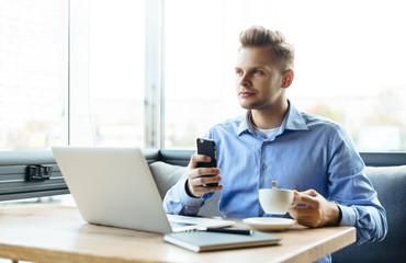 Young handsome man sitting in office with cup of coffee and working on project connected with modern cyber technologies. Businessman with notebook trying to keep deadline in digital marketing sphere.