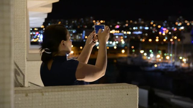 a woman stands on the balcony of the hotel and takes video a nigth city on the phone
