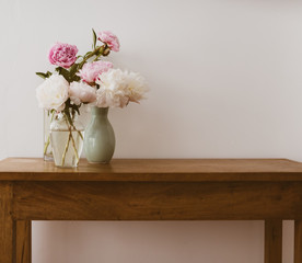 Pink and white peonies in glass and ceramic vases on oak wooden table against neutral wall - warm matte filter effect and selective focus