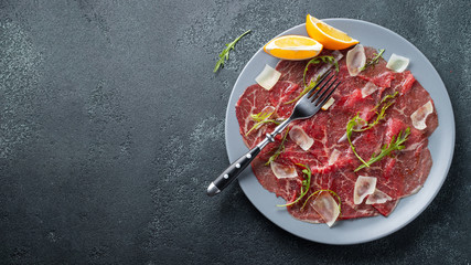 Marbled beef carpaccio with arugula, lemon and parmesan cheese on dark concrete table. Top view,...