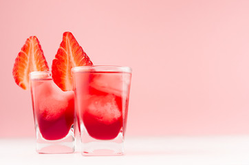Fresh juicy cocktails in two shot glasses with ice cubes, strawberry slice in modern youth interior in pastel soft pink color and white wood table.