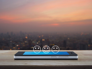 Excellent smiley face rating icon on modern smart mobile phone screen on wooden table over blur of cityscape on warm light sundown, Business customer service evaluation and feedback rating online conc