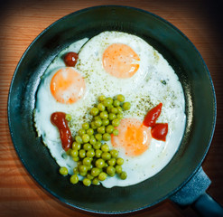 A frying pan with three fried eggs as well as peas and ketchup on a wooden background, top view.