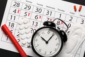 Menstrual calendar with pills, pad and alarm clock on dark background