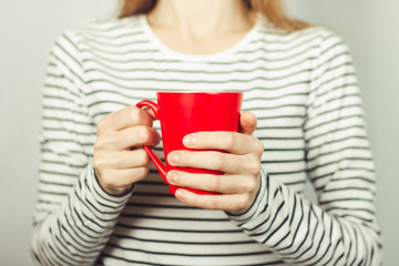 A girl in a striped T-shirt holds a red cup of coffee or tea in front of her. Breakfast concept, coffee break.