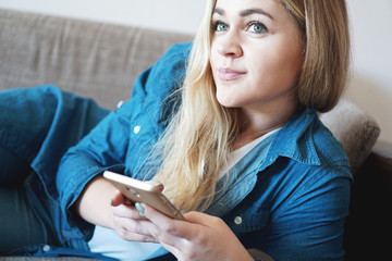 Portrait of dreamy thoughtful woman lying on couch pillow holding smart phone in hands looking away having fun pleasure harmony comfort