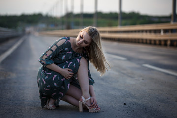 Young blonde girl on the road in the evening soft light of the sun. A girl in a green dress with long hair, the dress is blown by the wind.