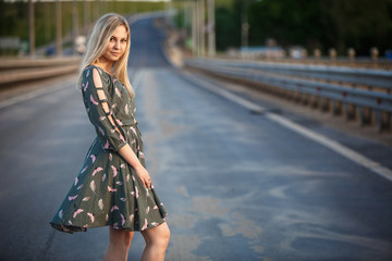 Young blonde girl on the road in the evening soft light of the sun. A girl in a green dress with...