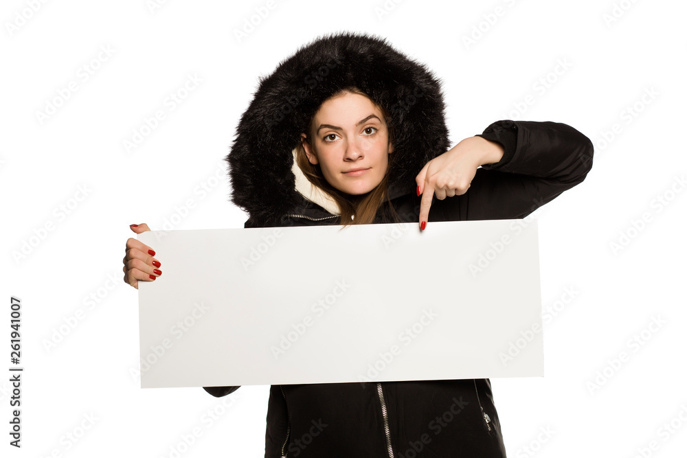 Wall mural Young model in winter jacket with hood shows an empty advertising board on white background - Wall murals