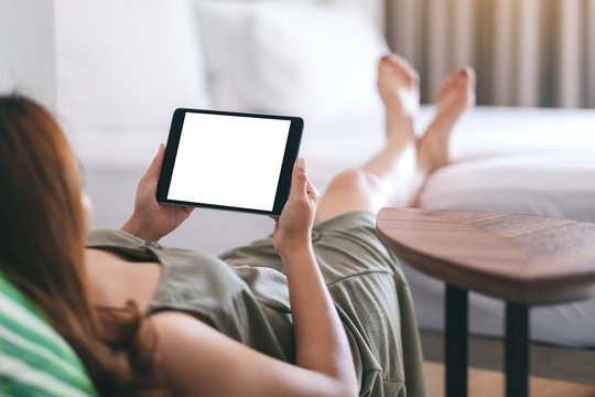 Mockup image of a woman holding and using tablet pc with blank screen horizontally while relaxing and lying on the bed