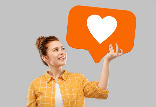 social media marketing, internet and people concept - smiling red haired teenage girl in checkered shirt holding heart icon over grey background