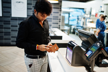 Indian man holding wallet behind his pay cash desk with order screen and card payment terminal in food cafe.