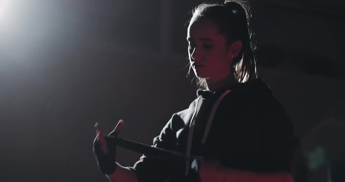 Young woman wrapping hands with black boxing wraps in the dark ring. Silhouette. Boxing concept