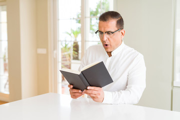 Middle age man reading a book at home wearing glasses scared in shock with a surprise face, afraid and excited with fear expression