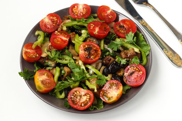Salad with aubergines and cherry tomatoes on a dark plate on a white background, top view, There is a knife and fork on the table