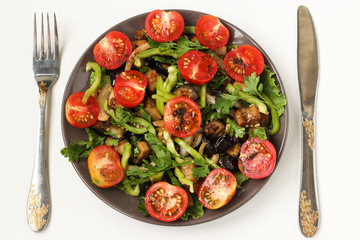 Salad with aubergines and cherry tomatoes on a dark plate on a white background, top view, There is a knife and fork on the table