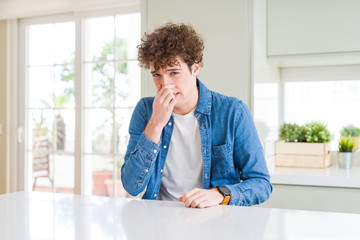 Young handsome man wearing casual denim jacket at home smelling something stinky and disgusting, intolerable smell, holding breath with fingers on nose. Bad smells concept.
