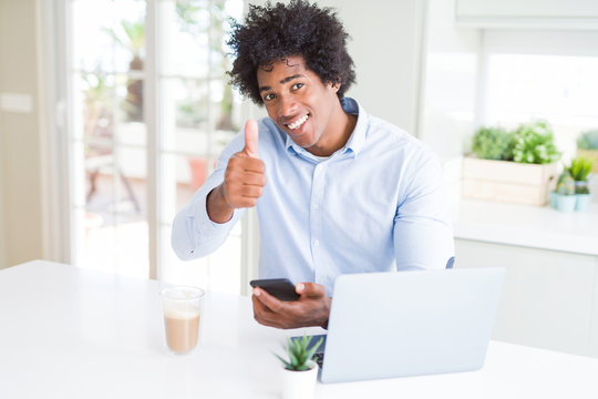 African American business man using smartphone and laptop happy with big smile doing ok sign, thumb up with fingers, excellent sign