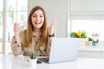 Beautiful young woman working with computer takes a break to drink glass of water very happy and excited, winner expression celebrating victory screaming with big smile and raised hands